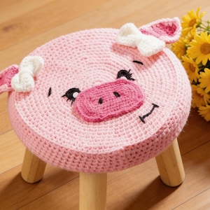 May include: A handmade, pink crochet pig stool with a round seat. The pig face features black eyes, a pink snout, and white bow accents on the ears. The stool has three wooden legs and is a charming piece of children's furniture.