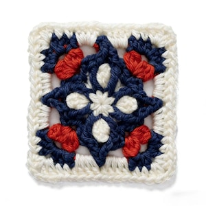 Puede incluir: Una pieza cuadrada de ganchillo con un borde crema y un diseño central en azul marino, blanco y rojo. El diseño presenta formas florales y patrones geométricos, mostrando una estética hecha a mano.