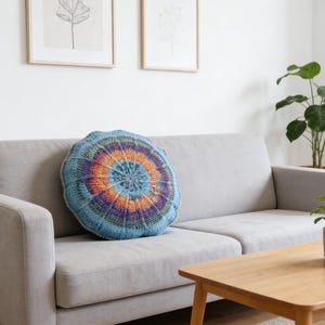 May include: A round, crocheted pillow with a vibrant color scheme of blue, orange, purple, and green. The pillow is placed on a light gray sofa in a living room setting. The room also features a wooden coffee table and potted plants.