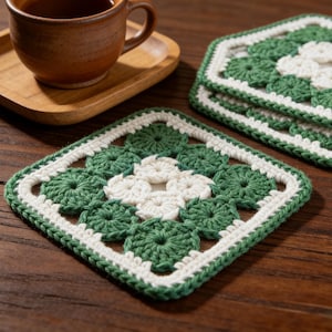 Può includere: Sottobicchieri quadrati all'uncinetto fatti a mano con un motivo floreale verde e bianco. I sottobicchieri sono impilati su una superficie di legno, con una tazza da tè marrone e un vassoio di legno sullo sfondo. Articolo decorativo.