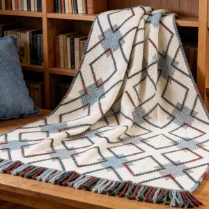 May include: Cream-colored knitted throw blanket with a geometric diamond pattern in blue, red, and green. The blanket has a fringed edge and is draped over a wooden surface. A denim pillow rests nearby.