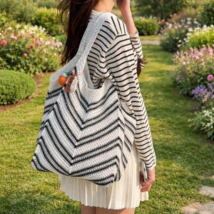 May include: A large, open-weave tote bag with a black and white chevron pattern. The bag has a wide strap and is adorned with a small, colorful charm. The person is wearing a striped long-sleeve shirt and a white skirt. The background is a garden.