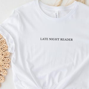 Könnte beinhalten: Ein weißes kurzärmeliges T-Shirt mit dem Text LATE NIGHT READER, der in schwarzen Großbuchstaben mittig auf der Brust aufgedruckt ist. Das Kleidungsstück hat einen Rundhalsausschnitt und eine Standardpassform mit schlichtem Design.