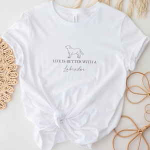 Könnte beinhalten: Ein weißes kurzärmeliges T-Shirt mit einer schwarzen Strichzeichnung eines Labrador-Hundes mittig auf der Brust. Unter dem Hund steht der Text "LIFE IS BETTER WITH A Labrador" in einer Kombination aus schwarzer Block- und Schreibschrift.