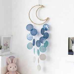 Puede incluir: Móvil decorativo con forma de media luna hecho de material tejido, con elementos circulares y ovalados azules y turquesas colgantes. El móvil está suspendido de una cuerda y cuelga frente a una pared blanca. Una decoración de guardería.