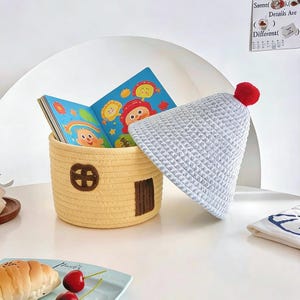 May include: A yellow woven storage basket shaped like a house, with a brown door and window, holds a colorful children's book. A light gray waffle-knit hat with a red pom-pom rests beside it. The scene is set on a white surface.