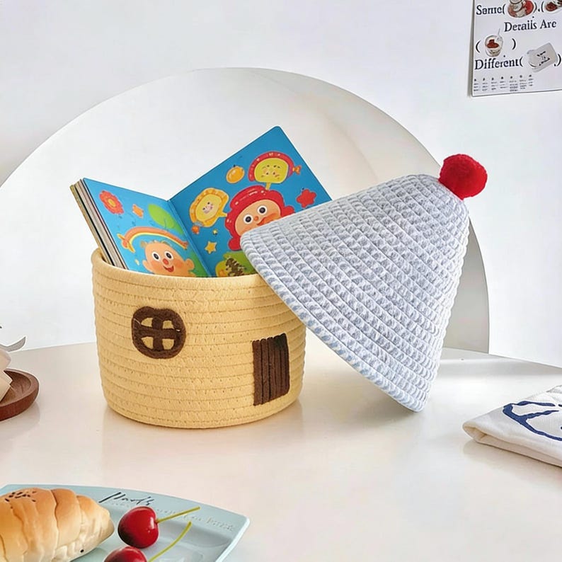 May include: A yellow woven storage basket shaped like a house, with a brown door and window, holds a colorful children's book. A light gray waffle-knit hat with a red pom-pom rests beside it. The scene is set on a white surface.