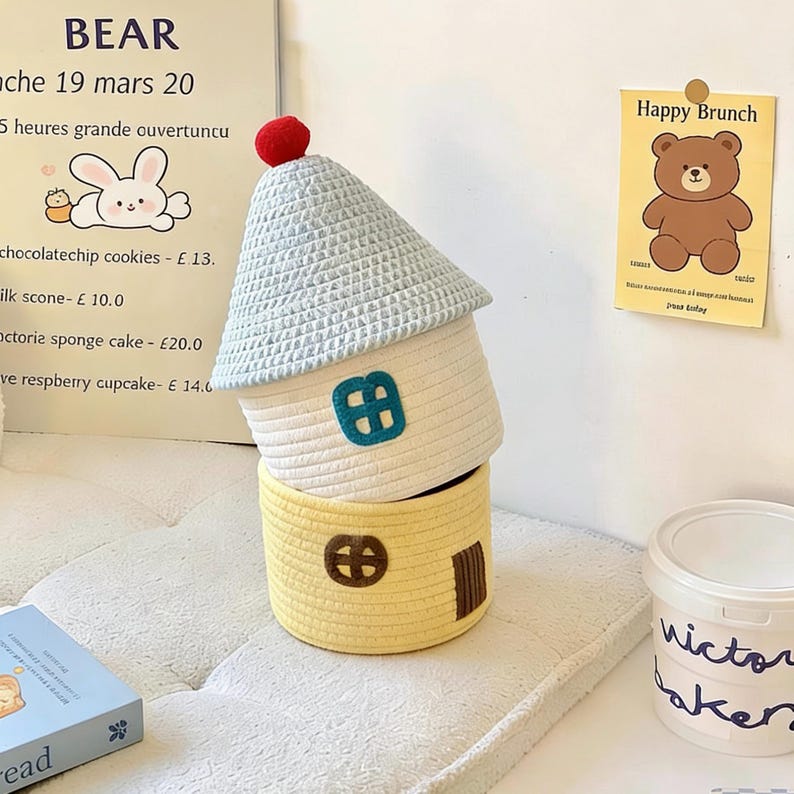 May include: Three woven storage baskets shaped like houses. The top basket has a light blue roof and a red pom-pom. The middle basket is white with a teal window. The bottom basket is yellow with a brown door and window.
