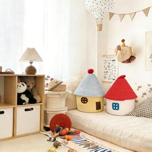 May include: Two house-shaped storage baskets with red and blue roofs. One is yellow with a blue roof, the other is white with a red roof. The baskets are on a white surface, with a rug and toys in the foreground.