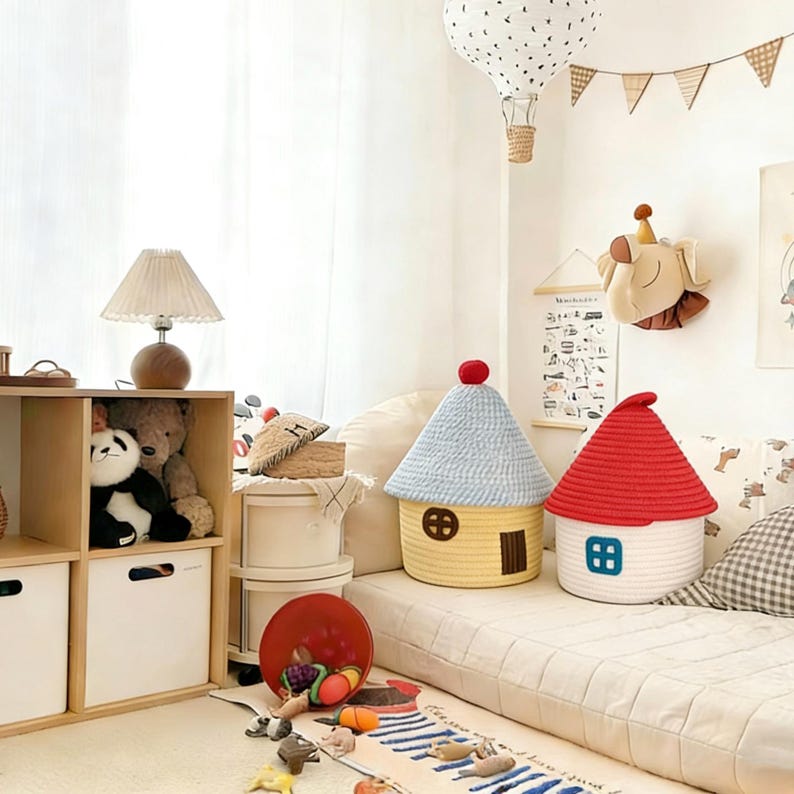 May include: Two house-shaped storage baskets with red and blue roofs. One is yellow with a blue roof, the other is white with a red roof. The baskets are on a white surface, with a rug and toys in the foreground.