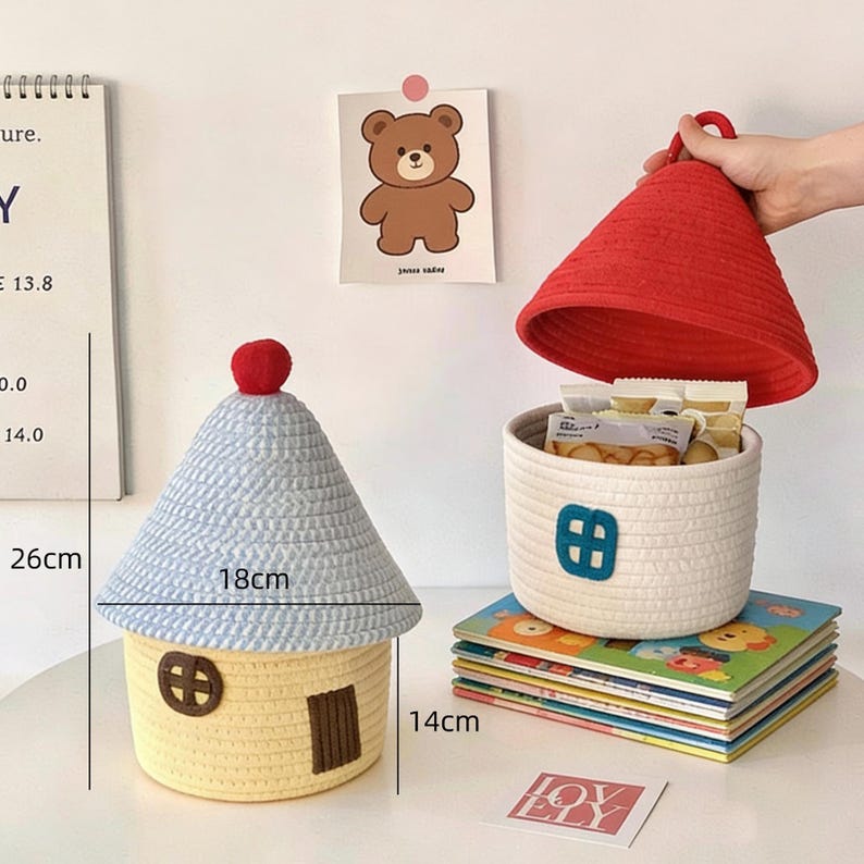 May include: A set of two decorative storage baskets. One is house-shaped, yellow with a blue roof, 26cm tall, 18cm wide, and 14cm deep. The other is a round white basket with a red lid. Both are made of woven material.