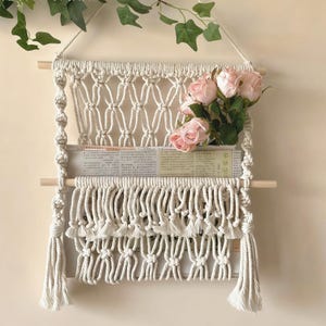 May include: Cream-colored macrame wall hanging with a wooden dowel and tassels. The wall decor features a woven design with a pocket holding a newspaper and a bouquet of pink roses.