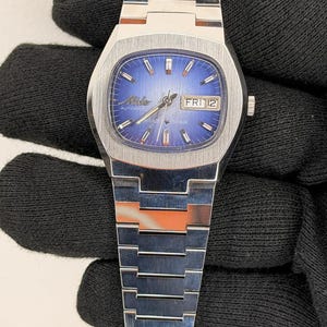 Puede incluir: Un reloj de pulsera automático Mido Multi Star vintage. El reloj presenta una esfera azul con agujas e indicadores plateados, y una pantalla de día y fecha. La caja y el brazalete de acero inoxidable tienen un diseño geométrico. El reloj se muestra sobre un fondo negro.