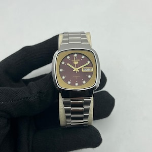 Könnte beinhalten: Vintage Seiko Automatik-Uhr mit rechteckigem Edelstahlgehäuse und braunem Zifferblatt. Die Uhr verfügt über goldfarbene Akzente, Diamant-Stundenmarkierungen und eine Tagesdatumsanzeige. Edelstahlarmband.