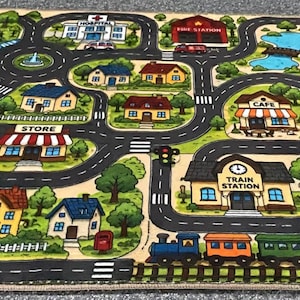 May include: A rectangular play rug featuring a colorful illustrated town map with roads, a train track, and various buildings including a school, hospital, fire station, store, cafe, and train station, surrounded by green trees and landscaping.