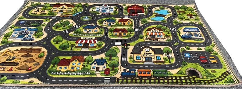 May include: A rectangular play rug featuring a colorful illustrated town map with roads, a train track, and various buildings including a school, hospital, fire station, store, cafe, and train station, surrounded by green trees and landscaping.