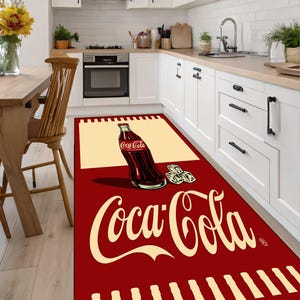 Könnte beinhalten: Ein rechteckiger Küchenläufer mit tiefrotem Hintergrund und einem großen cremefarbenen Coca-Cola-Logo. Das Design zeigt eine grafische Illustration einer Coca-Cola-Glasflasche und Eiswürfeln sowie cremefarbene Akzente am Rand.
