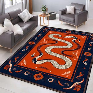 Puede incluir: Una alfombra rectangular que presenta una gran ilustración de una serpiente sinuosa en blanco y negro sobre un fondo naranja vibrante. La alfombra está enmarcada por un borde ancho de color azul oscuro decorado con motivos florales.