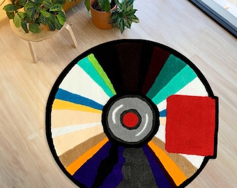 May include: A circular tufted rug featuring a radial design of multicoloured segments in shades of teal, yellow, orange, purple, and black, centred with a grey and red circular motif and a prominent solid red rectangular patch on the right side.
