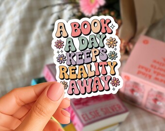 Op de afbeelding: Een kleurrijke sticker met de tekst "A BOOK A DAY KEEPS REALITY AWAY" in een retro lettertype. De sticker heeft pastelkleuren en bloemmotieven. De sticker wordt door een hand vastgehouden, met boeken en bloemen op de achtergrond.