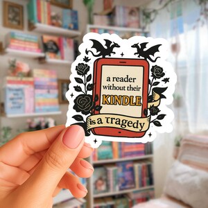 Op de afbeelding: Een witte sticker met een rood tabletontwerp, zwarte draken en rozen. De sticker heeft de tekst "a reader without their KINDLE is a tragedy" in zwart en beige. De sticker wordt in een hand gehouden.