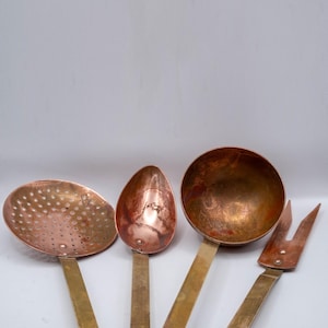 May include: A collection of four copper kitchen utensils: a slotted spoon, a solid spoon, a ladle, and a fork. The copper tools have brass handles and are arranged against a white background. These vintage cooking tools are ideal for a rustic kitchen.