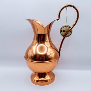 May include: A copper-colored pitcher with a curved handle and a flared spout. The pitcher has a round body and a pedestal base. A small tag hangs from the handle. The pitcher is likely used for serving beverages.