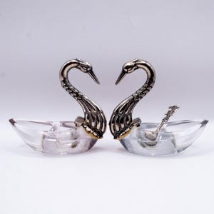 May include: Two swan-shaped salt cellars. Each has a clear glass bowl and a silver-coloured swan head. One cellar contains a small spoon. The swans face each other, creating a decorative display.