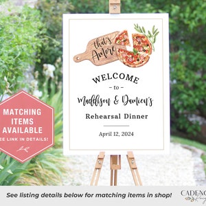 May include: A white sign with a gold border announcing a rehearsal dinner for Maddison & Damien's. The sign features a watercolor illustration of a pizza with the text "that's Amore" on a wooden cutting board. The date of the dinner is April 12, 2024.