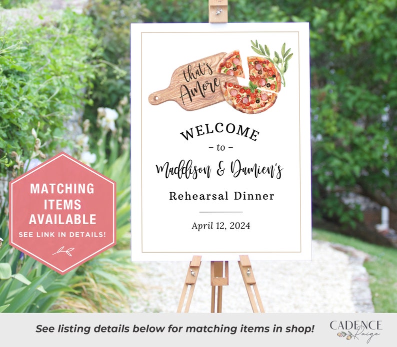 May include: A white sign with a gold border announcing a rehearsal dinner for Maddison & Damien's. The sign features a watercolor illustration of a pizza with the text "that's Amore" on a wooden cutting board. The date of the dinner is April 12, 2024.