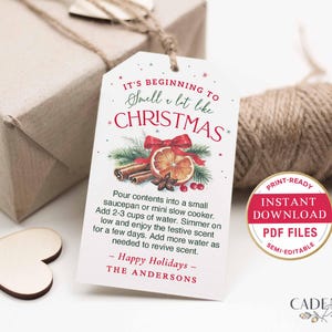 Puede incluir: Una etiqueta de regalo blanca con el texto "It's beginning to smell a lot like Christmas" e instrucciones para una olla a fuego lento. La etiqueta presenta una ilustraci&oacute;n de acuarela de palitos de canela, una rodaja de naranja y un lazo rojo. Una caja de regalo y cuerda est&aacute;n en el fondo.