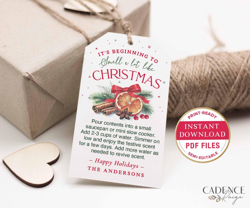 Puede incluir: Una etiqueta de regalo blanca con el texto "It's beginning to smell a lot like Christmas" e instrucciones para una olla a fuego lento. La etiqueta presenta una ilustraci&oacute;n de acuarela de palitos de canela, una rodaja de naranja y un lazo rojo. Una caja de regalo y cuerda est&aacute;n en el fondo.