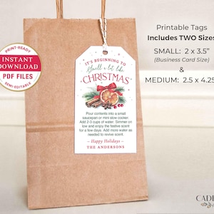 Puede incluir: Una bolsa de regalo de papel marr&oacute;n con una etiqueta blanca que dice "It's beginning to smell a bit like Christmas". La etiqueta presenta un dise&ntilde;o de acuarela de palitos de canela, una rodaja de naranja y un lazo rojo. La etiqueta est&aacute; atada a la bolsa con hilo.