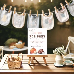 K&ouml;nnte beinhalten: Ein wei&szlig;es Schild mit braunem Text, auf dem "Bibs for Baby" steht. Das Schild steht auf einem Holzstaffelei und hat ein Herbstthema mit K&uuml;rbissen und einem Fu&szlig;ball. Das Schild steht auf einem Holztisch mit einem Weidenkorb, einer Vase mit wei&szlig;en Blumen und zwei kleinen wei&szlig;en Tassen.