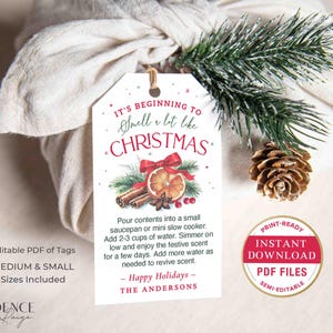 Puede incluir: Una etiqueta de regalo blanca con el texto "It's beginning to smell a lot like Christmas" en rojo y verde. La etiqueta presenta un dise&ntilde;o festivo con un lazo, una rodaja de naranja, palitos de canela y ramitas de pino.