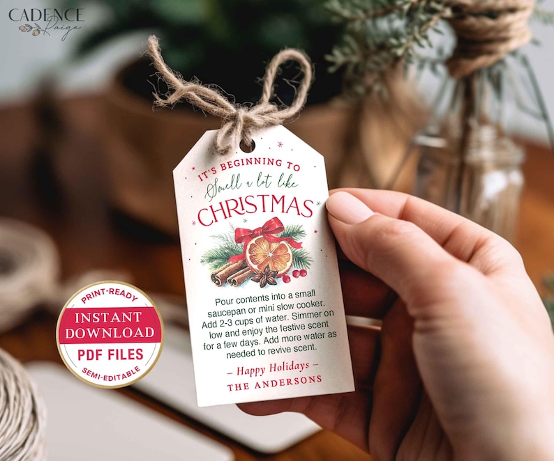 Puede incluir: Una etiqueta de regalo blanca con tem&aacute;tica navide&ntilde;a con el texto "It's Beginning to Smell a lot like Christmas". La etiqueta presenta ilustraciones de palitos de canela, una rodaja de naranja y un lazo rojo. La etiqueta est&aacute; atada con hilo.