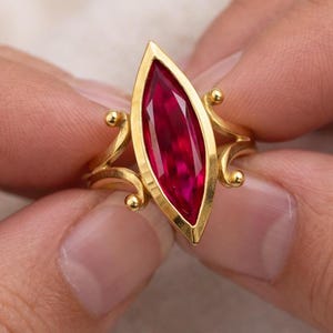 以下が含まれることがあります： 深紅色のマーキス型宝石をあしらったゴールドリング。リングは、装飾的な渦巻きと小さな丸いアクセントが特徴的なユニークなデザインです。宝石はゴールドベゼルにセットされ、印象的なコントラストを生み出しています。