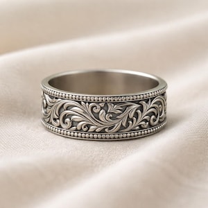 Può includere: Un anello d'argento con un intricato motivo floreale. L'anello presenta un design dettagliato di foglie e viticci a spirale, bordato da un bordo perlato. L'anello misura circa 0,8 cm di larghezza.