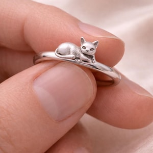Op de afbeelding: Zilveren ring met een gedetailleerde kat die op de band ligt. De kat is realistisch vormgegeven. De ring wordt door een persoon vastgehouden.