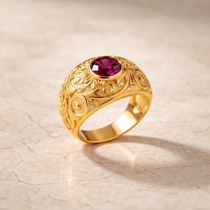 Könnte beinhalten: Ein goldener Ring mit einem ovalen, tiefroten Edelstein in der Mitte. Der Ring hat aufwendige Blumen- und Scroll-Designs mit kleinen, klaren Akzentsteinen. Das Band ist breit und poliert.