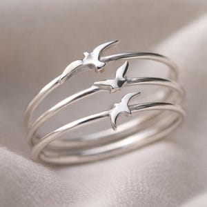 May include: A silver ring set featuring three bands, each adorned with a stylized bird design. The birds are evenly spaced around the ring, creating a layered effect. The ring is made of a shiny, reflective metal.