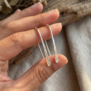 Può includere: Un paio di orecchini color argento con finitura martellata. Gli orecchini hanno una semplice forma a U allungata con una superficie strutturata. Gli orecchini sono tenuti tra le dita su uno sfondo neutro.