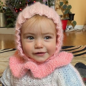 Könnte beinhalten: Ein kleines Kind trägt eine rosa gestrickte Mütze und einen passenden Pullover mit einem Rüschenkragen. Die Mütze hat eine Kapuze und der Pullover ist hellrosa und weiß.