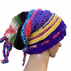 Könnte beinhalten: Ein handgefertigter, mehrfarbiger Strickhut mit langem, lockerem Design. Der Hut hat eine Mischung aus leuchtenden Farben, darunter Lila, Gelb, Rosa, Blau und Grün. Geflochtene Quasten hängen an den Seiten. Ein einzigartiges Accessoire.
