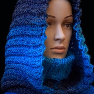 May include: A knitted hood and cowl set in shades of blue. The hood features a ribbed texture and a ruffled edge, while the cowl has a turtleneck design. The set is displayed on a mannequin, showcasing the handmade craftsmanship.