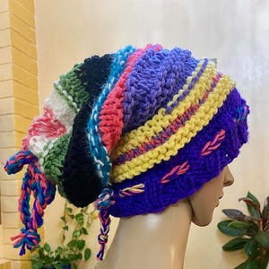 May include: A colourful crocheted beanie hat with a long, dangling tail. The hat is made of multiple colours of yarn, including purple, yellow, pink, green, and black.