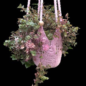 Peut inclure: Une plante suspendue dans un porte-plante macramé rose tricoté. La plante a des feuilles vertes et roses, avec quelques feuilles en cascade. Le porte-macramé est suspendu par des cordons roses et une perle en bois. Le fond est noir.