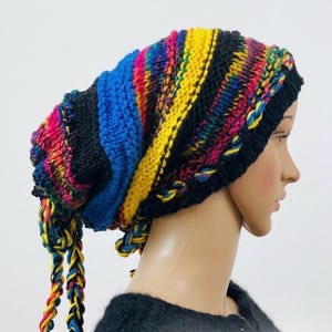 May include: A slouchy knit beanie featuring horizontal stripes in black, bright blue, yellow, and a multi-colored variegated yarn. The hat has a black ribbed brim and two braided tassels hanging from the back, crafted from the same colorful yarn.