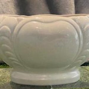 May include: Cream ceramic planter with two loop handles. The planter features an embossed floral design on the front and sides. It sits on a small, round base. The planter is displayed on a clear surface.
