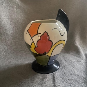 May include: A decorative ceramic vase featuring a black base and a curved black handle. The vase is adorned with abstract patterns in orange, red, yellow, green, and black on a cream backdrop. The vase has a rounded form.